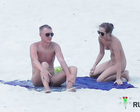 Two Nudists Settling onto a Blue Towel on White Sand