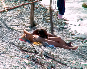 Nudists in foreplay on a towel at a sunlit pebble shoreline