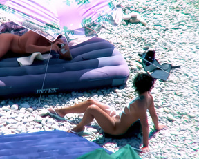 Two nudists resting on a sunlit pebble nude beach beside striped shade