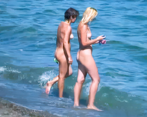 Two nudists wading through sunlit shallows at a sandy nude beach