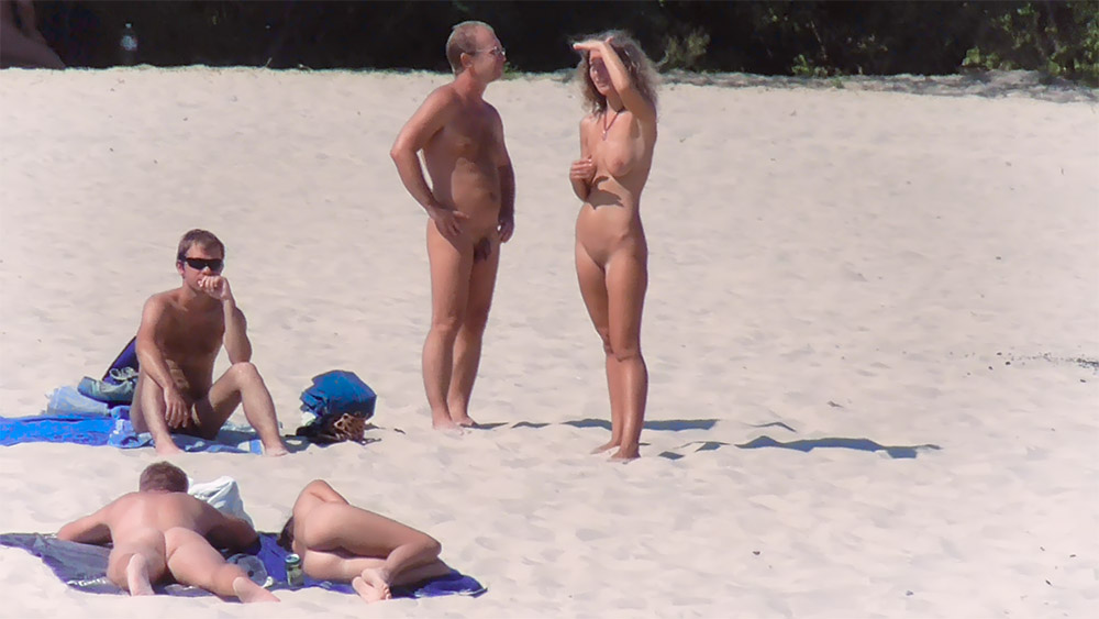 Dva nudisté stojící na písku u modré tašky