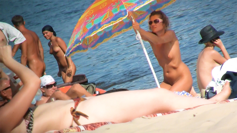 Two Nudists Setting Up a Colorful Umbrella by the Sea