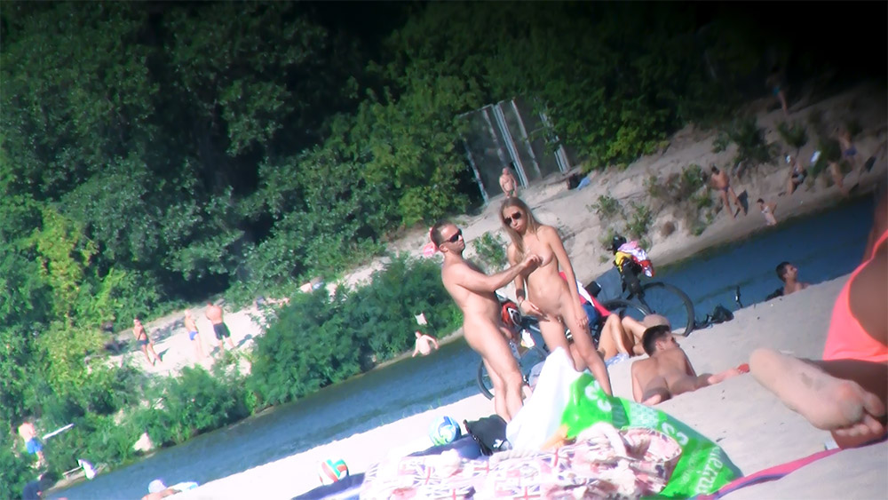 Nudisté stojí a povídají si na písčitém břehu řeky
