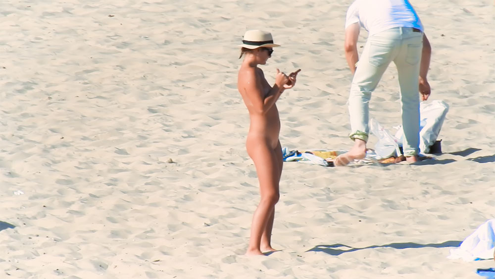 Olhando o celular numa praia de nudismo com um chapéu de palha