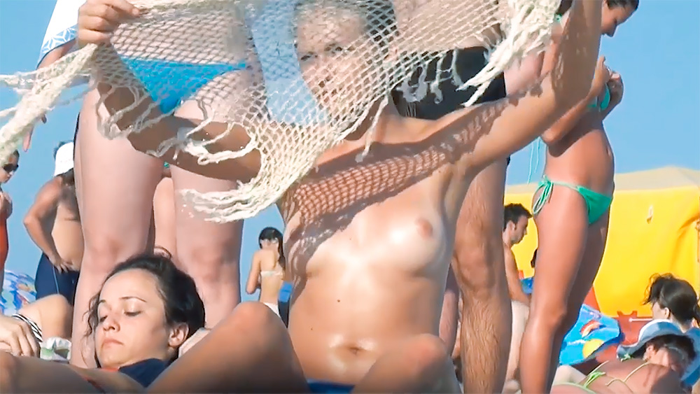 Two Topless Beach Sunbathers Beside a White Net