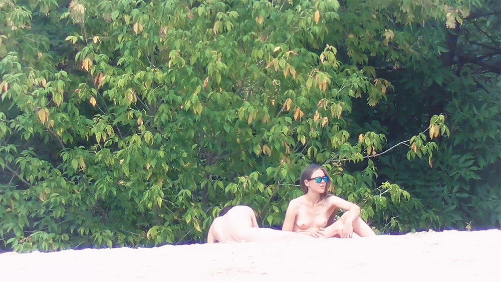 Naturistka na břehu řeky v zrcadlových slunečních brýlích opírající se dozadu