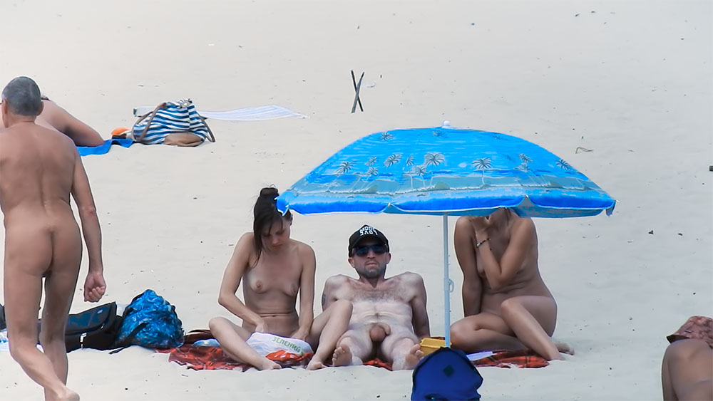 Three Nudists Sitting Under a Blue Palm-Print Beach Umbrella