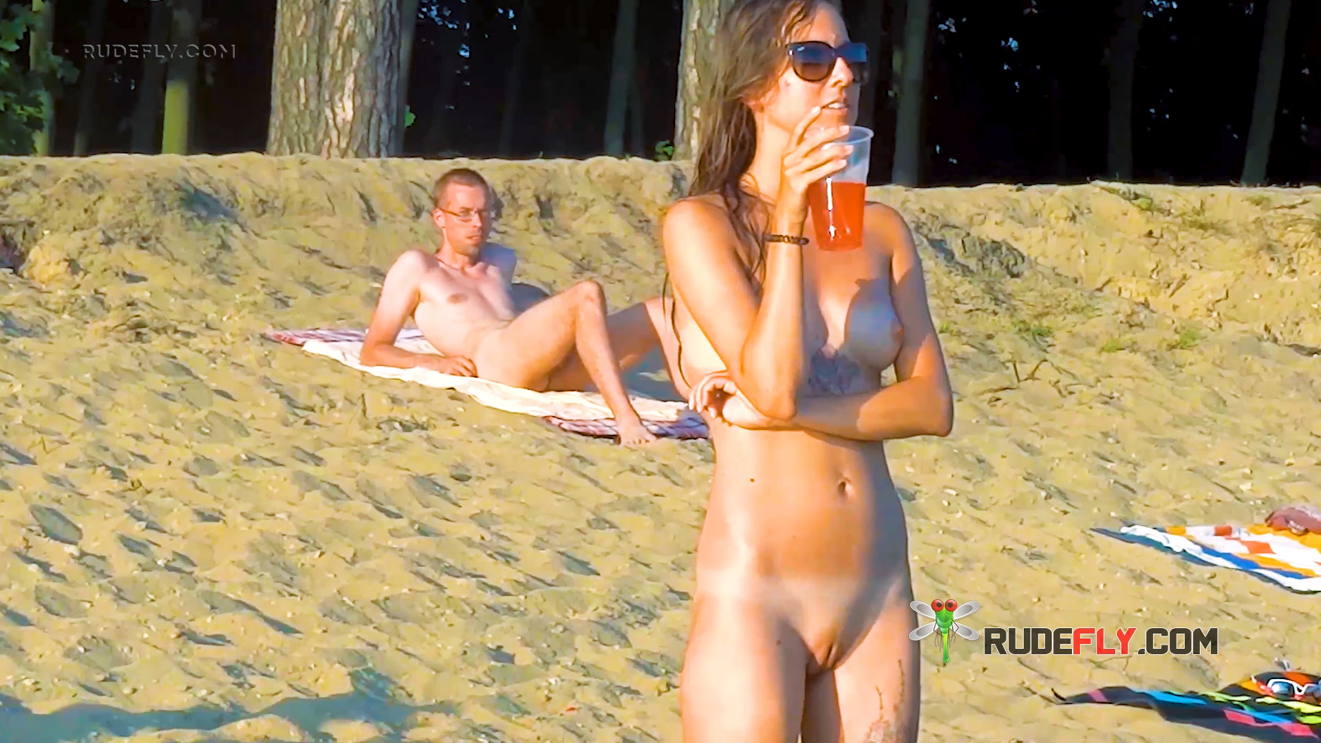 Two Nudists Standing With Orange Drinks on the Sand