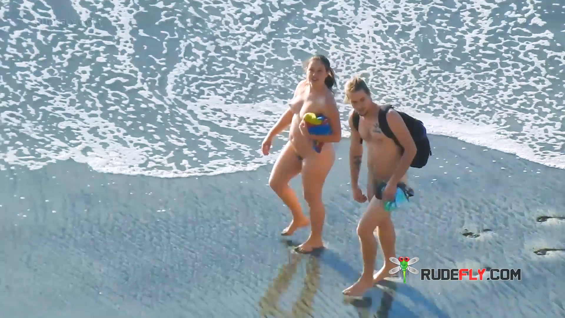 Two Nudists Strolling Past the Surf with Beach Gear