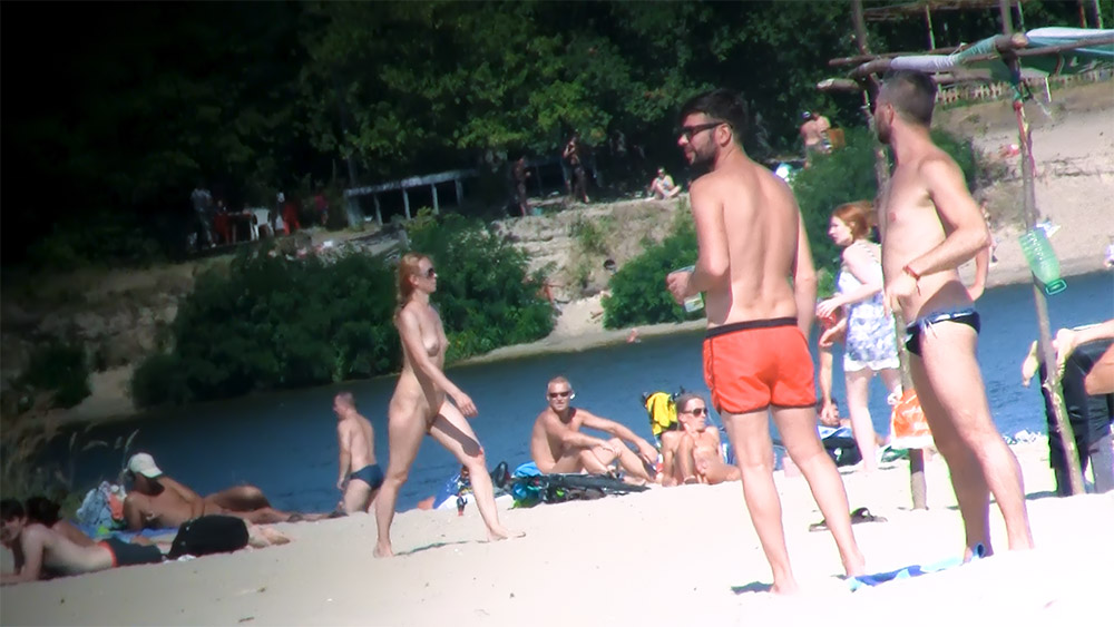 Štíhlá nahá žena procházející se mezi nudisty na břehu řeky