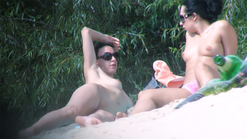 Two Nude Beach Sunbathers Resting Beside Leafy Bushes