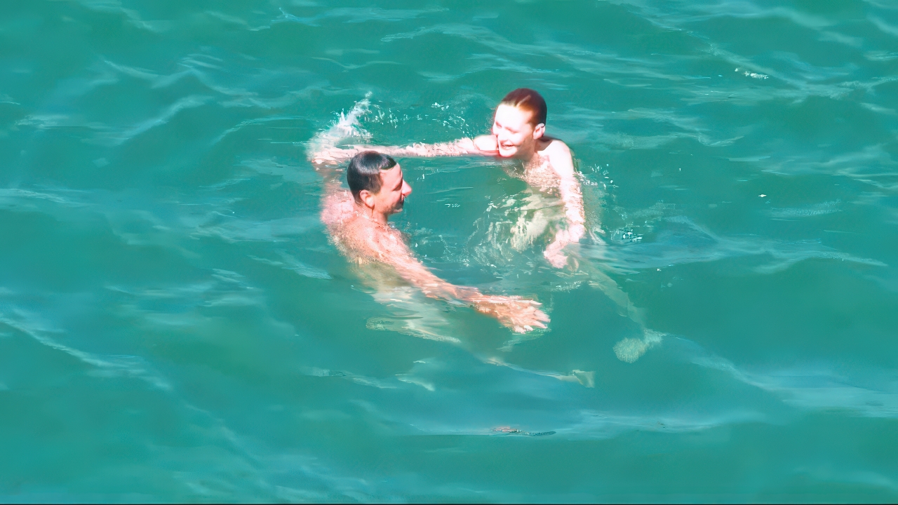 Due naturisti ridono nuotando nell'acqua turchese al sole