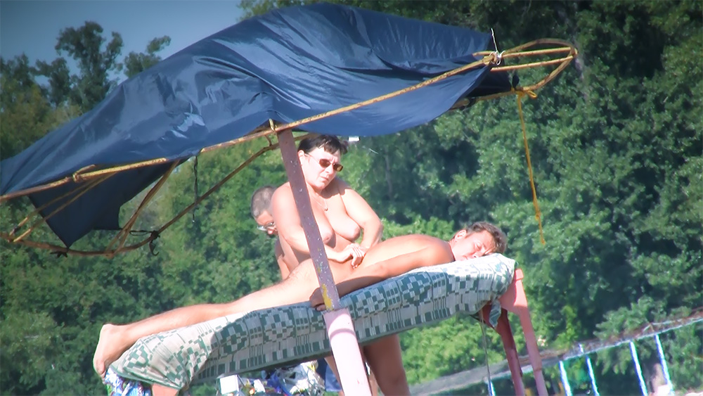 Three Naturists Sharing Shade on a Riverside Mattress