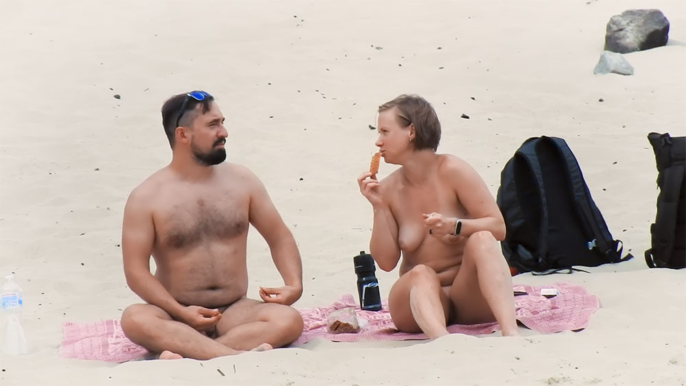 Two Nudists Sharing Snacks on a Pink Beach Towel