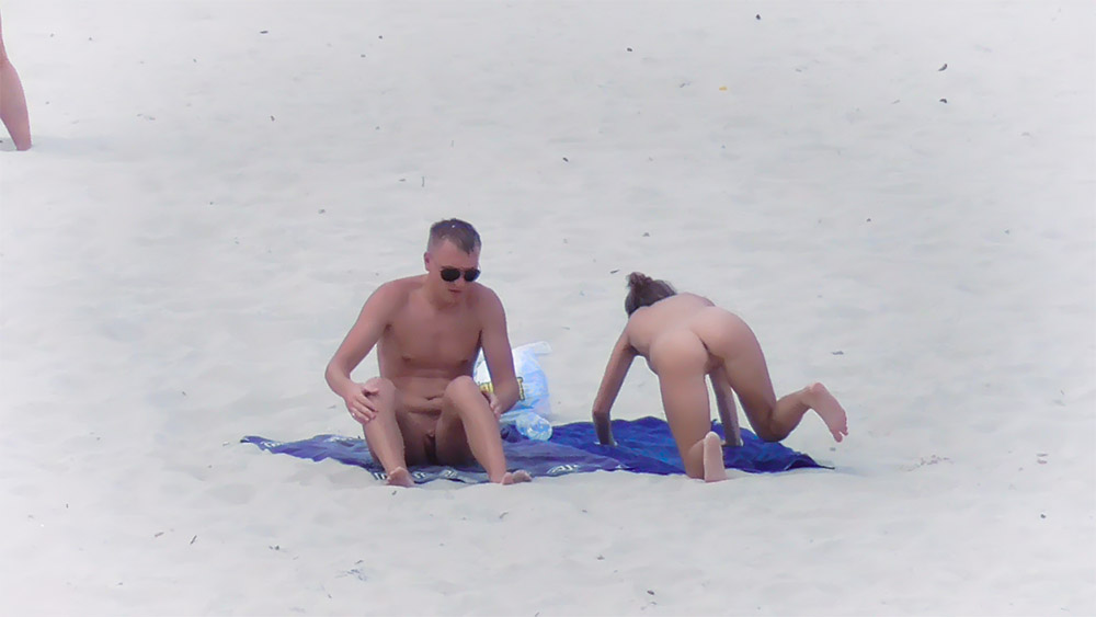 Two Nudists Settling onto a Blue Towel on White Sand