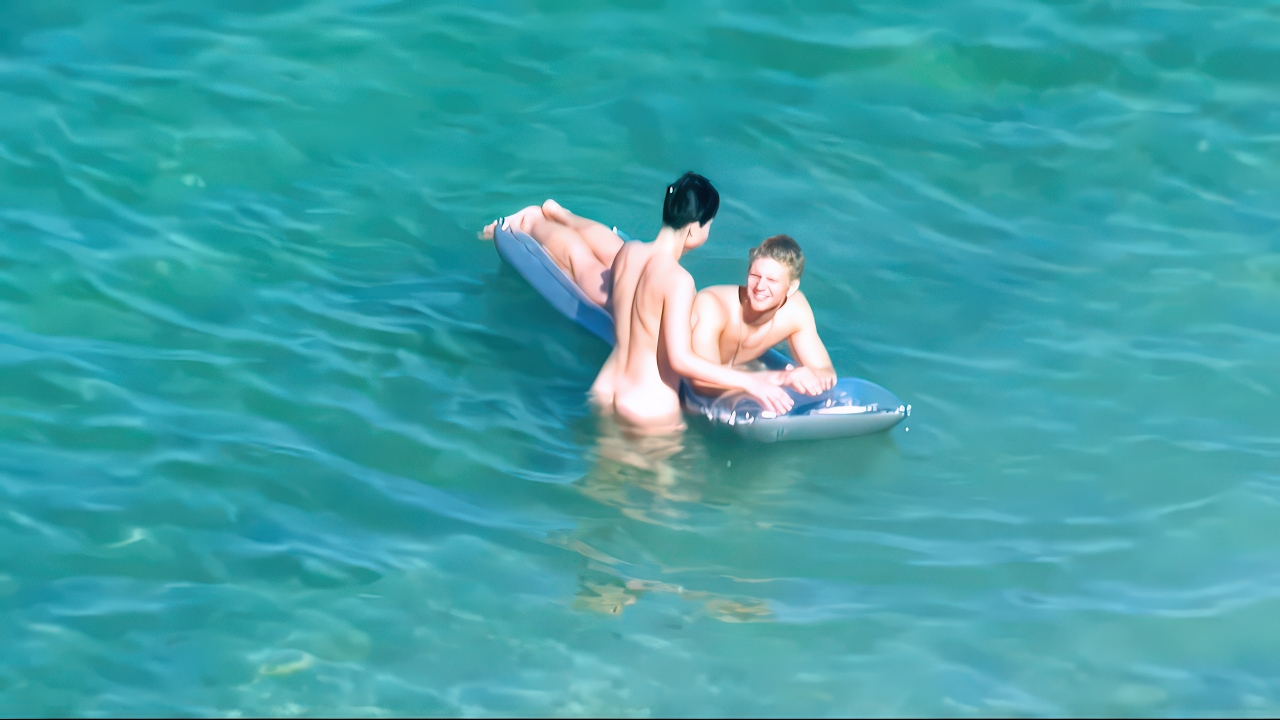 Dva nudisté odpočívají na modrém pontonu ve slunečné tyrkysové vodě