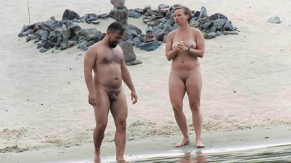 Two Nudists Pausing Near Stacked Stones on Sand