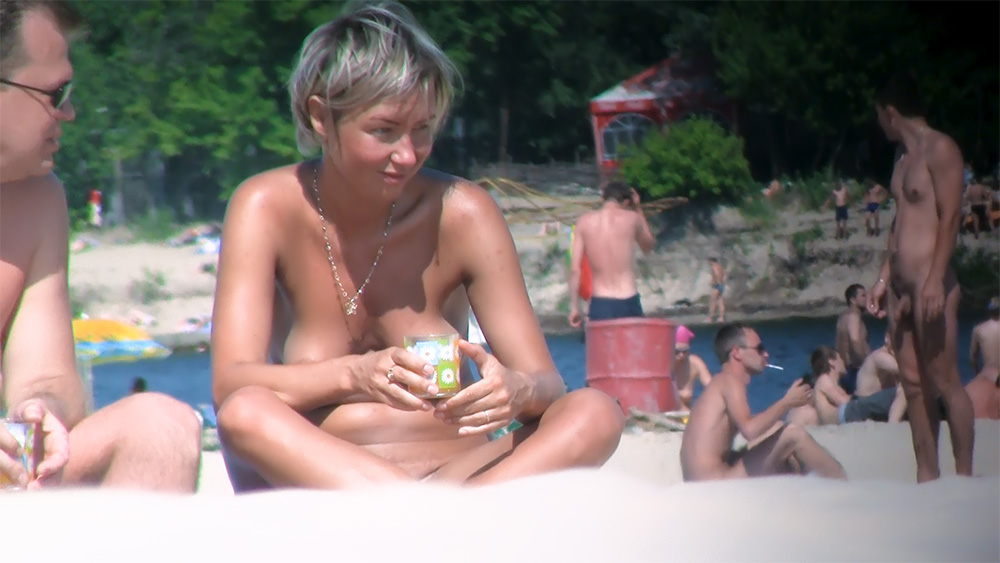 Tenant une boisson, assise sur une plage naturiste