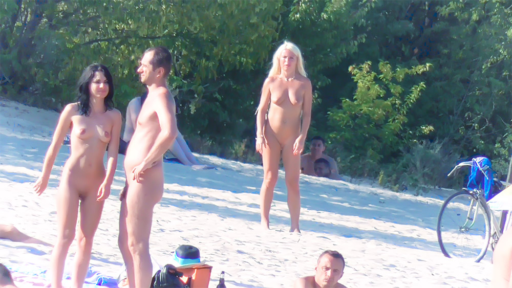 Naturistická pláž lemovaná stromy se třemi stojícími koupajícími