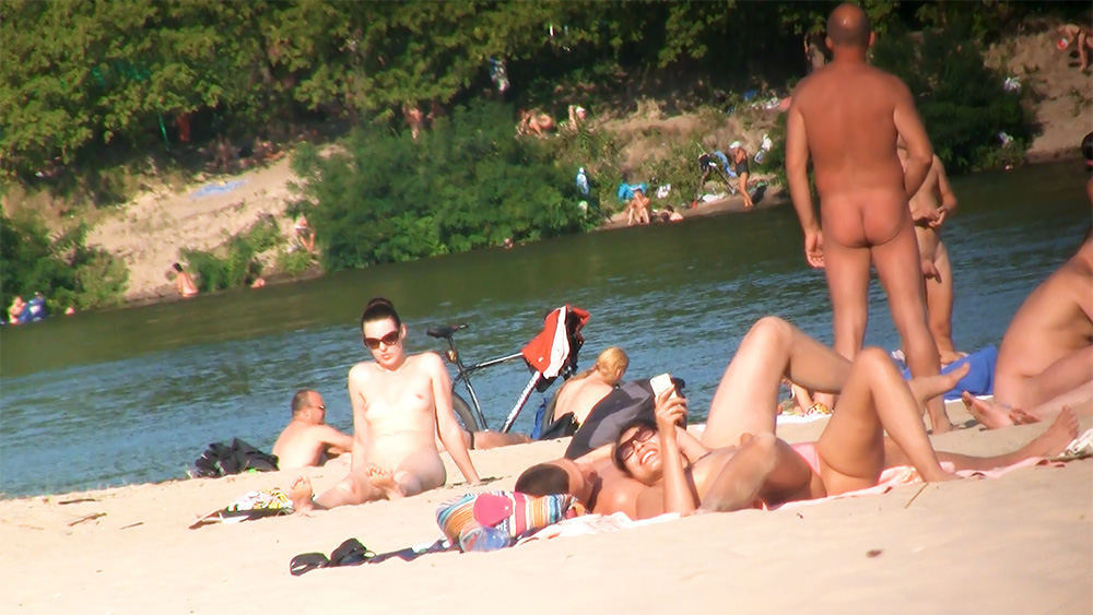 Five Naturists Gathered on Sand Beside Calm River Water