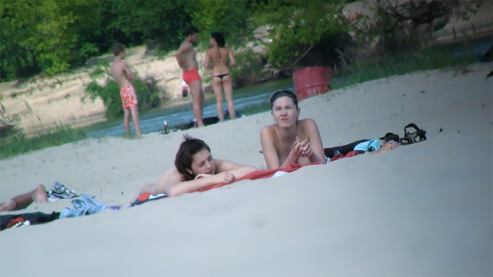 Two Women Sunbathing Topless on a Sandy Riverbank