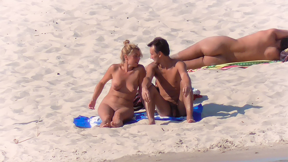 Tři naturisté odpočívající na ručnících na písečném břehu