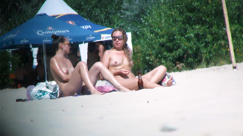 Two Nudists Sitting on Sand Beside a Blue Umbrella