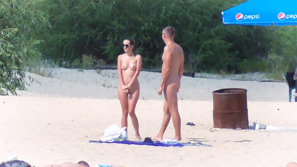 Tři nudisté stojící pospolu u rezavého sudu na pláži