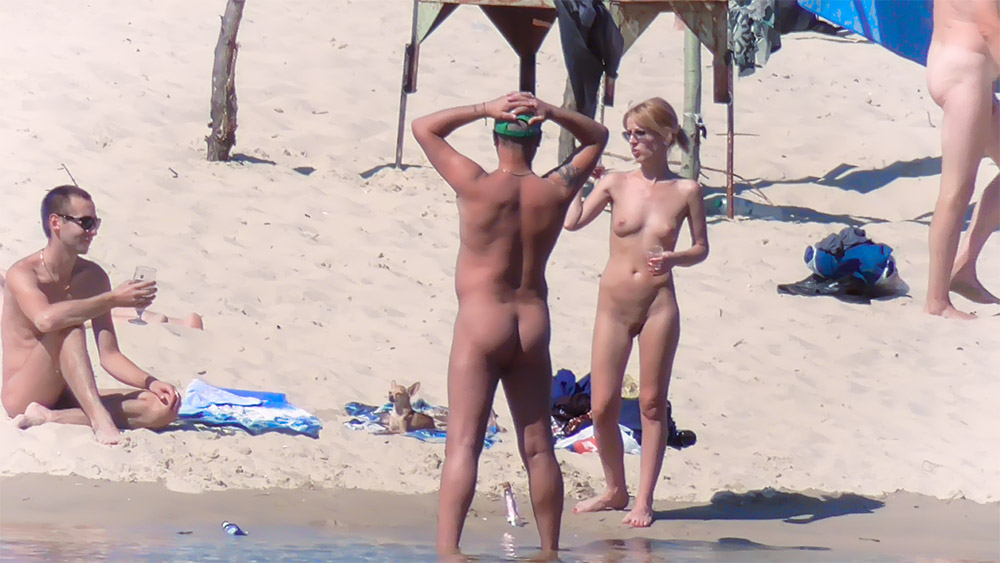 Two Nudists Conversing Near a Wooden Beach Shelter