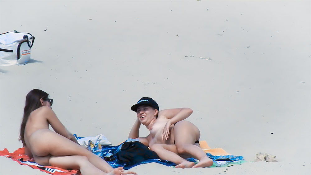 Two Nudists Lounging on Towels Across the Sand