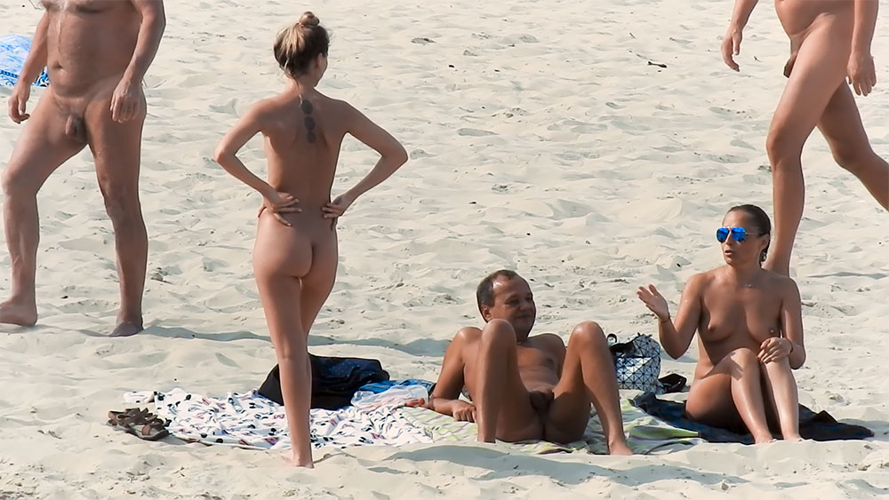 Cinco nudistas de pie y sentados juntos en la arena