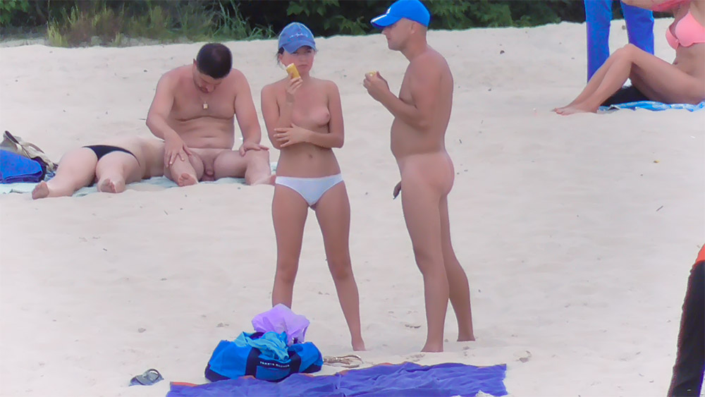 Drei Naturisten stehen mit blauen Kappen im Sand
